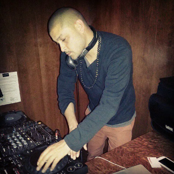 DJ CMAN leaning over the decks during the Grain Bar residency, Four Seasons Hotel Sydney, headphones and beads around the neck