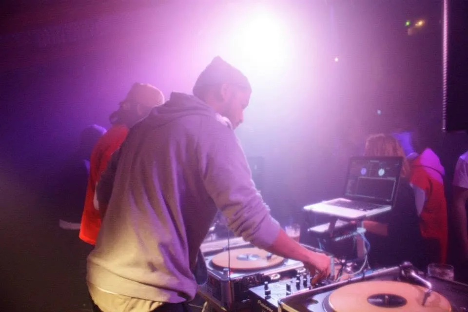 DJ CMAN leans into the mixer mid-blend, JuzzLikeDat beside him in the haze.